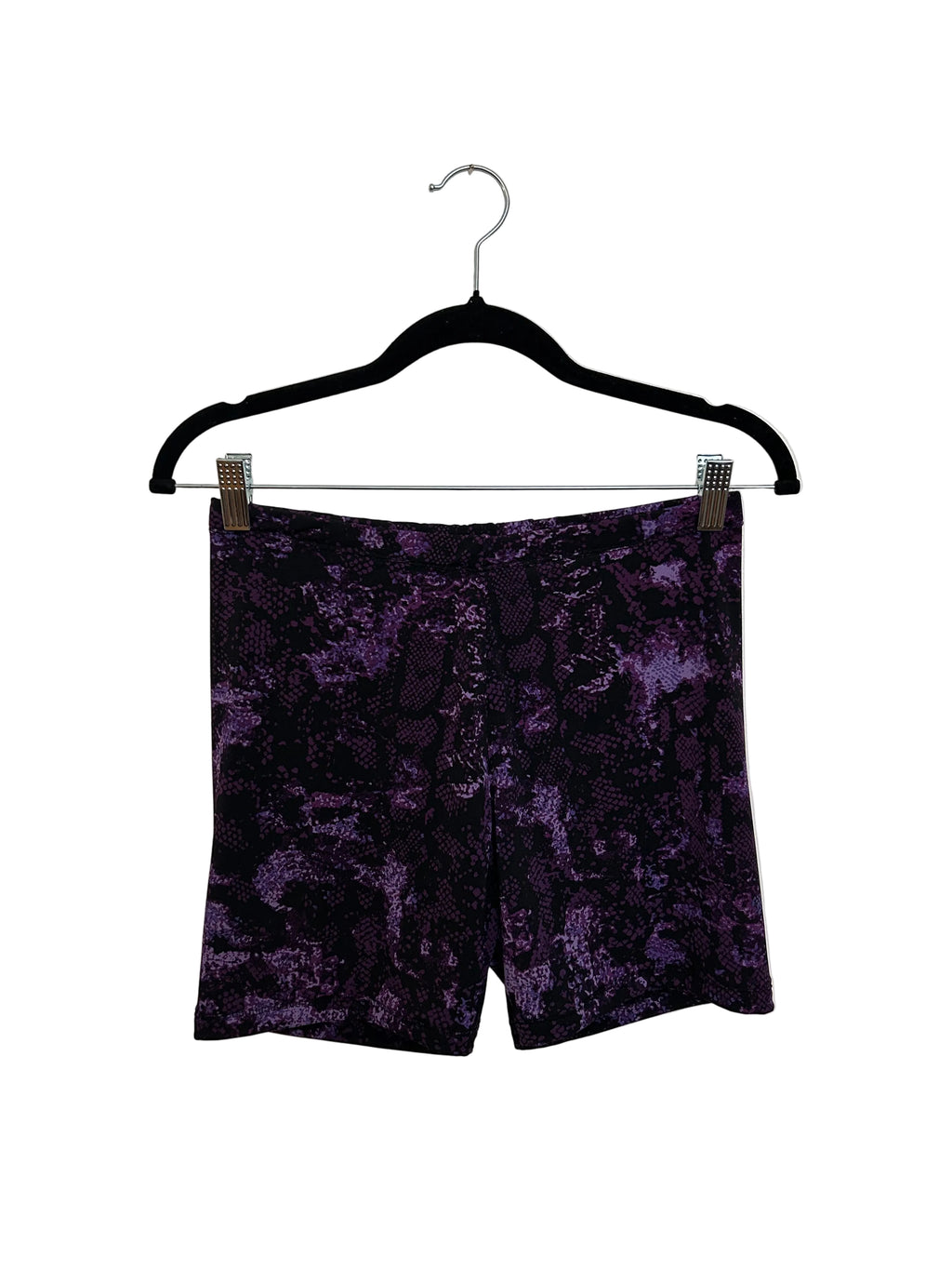 A pair of purple and black patterned shorts laid flat on a light beige surface, held in place by a black clothes hanger with metal clips.