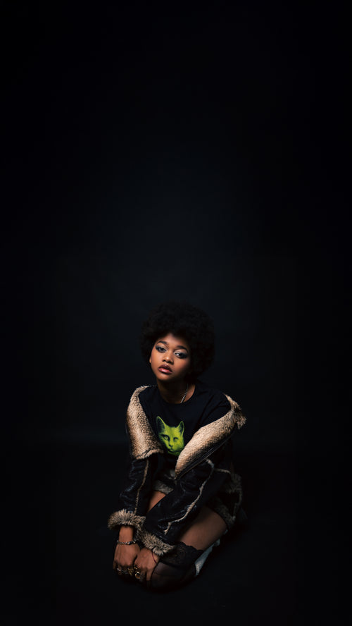 person sitting on a dark background wearing a patterned outfit with a green cat on her shirt.