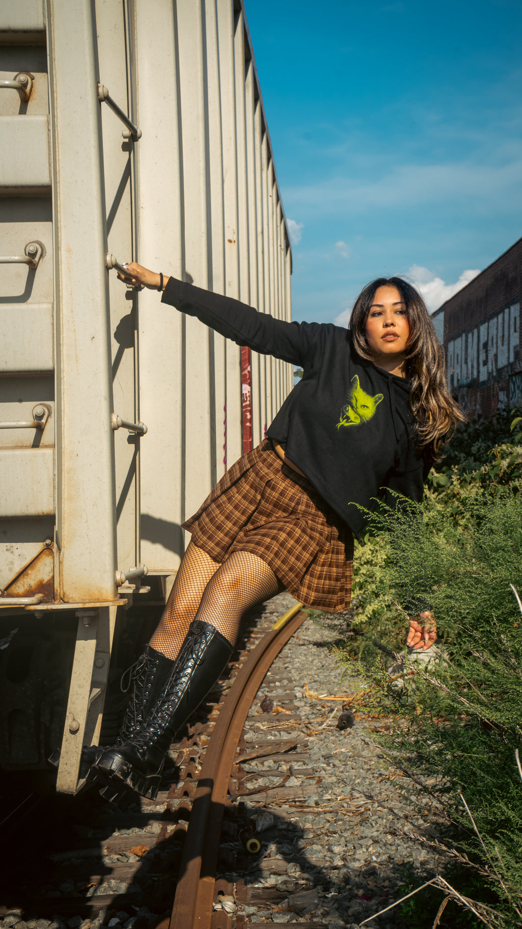 Model wearing Women's Cat Venom cropped hoodie – black cropped hoodie with neon green cat graphic, leaning against train car on railway track under blue sky, gothic streetwear style