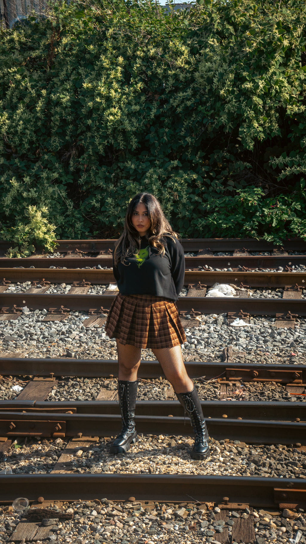 Person standing on train tracks with greenery in the backgroundModel wearing Women's Cat Venom cropped hoodie – black cropped hoodie with neon green cat graphic paired with plaid skirt, standing on train tracks with forest background, gothic streetwear style