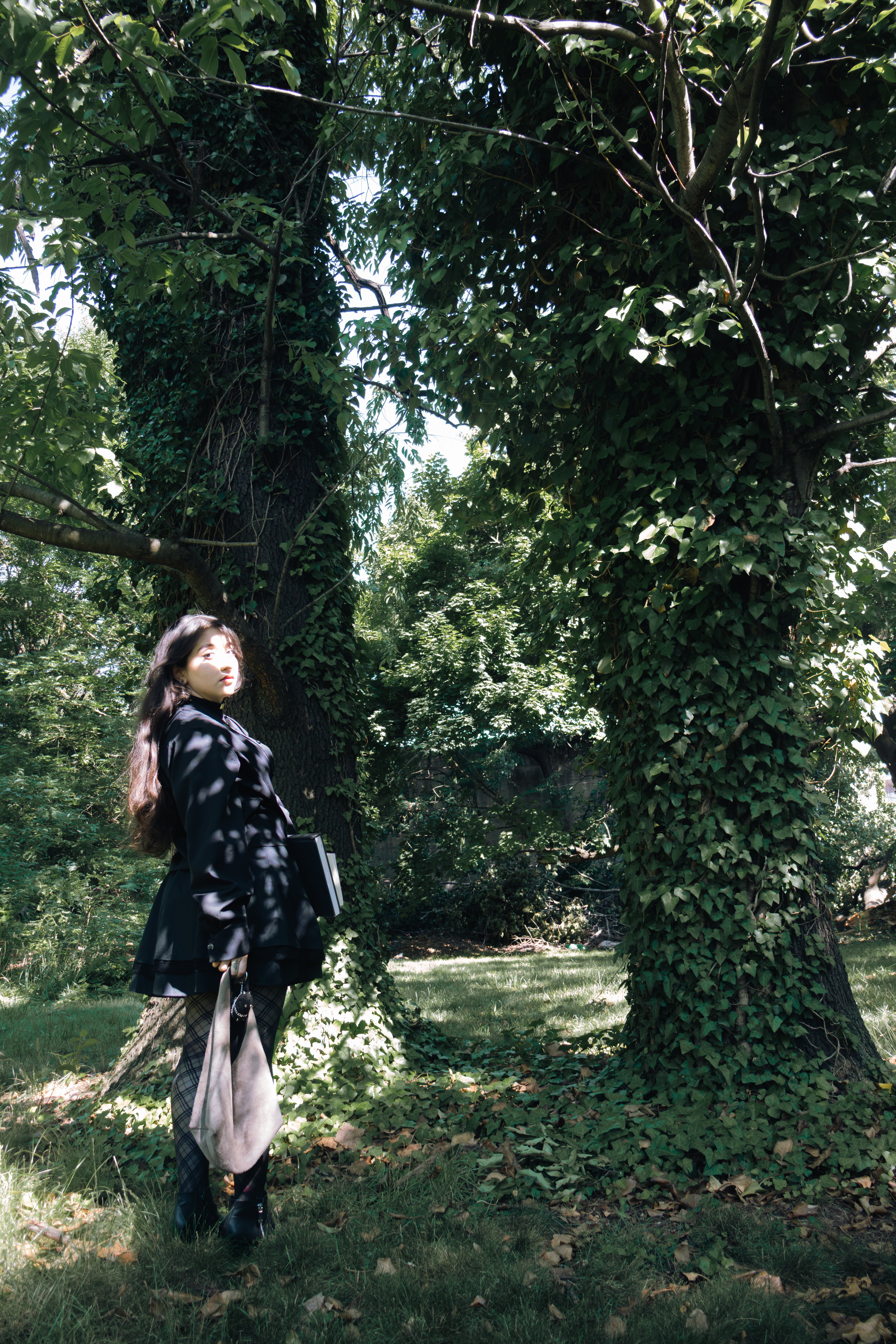 Person standing between two large trees covered in green ivy in a forest.