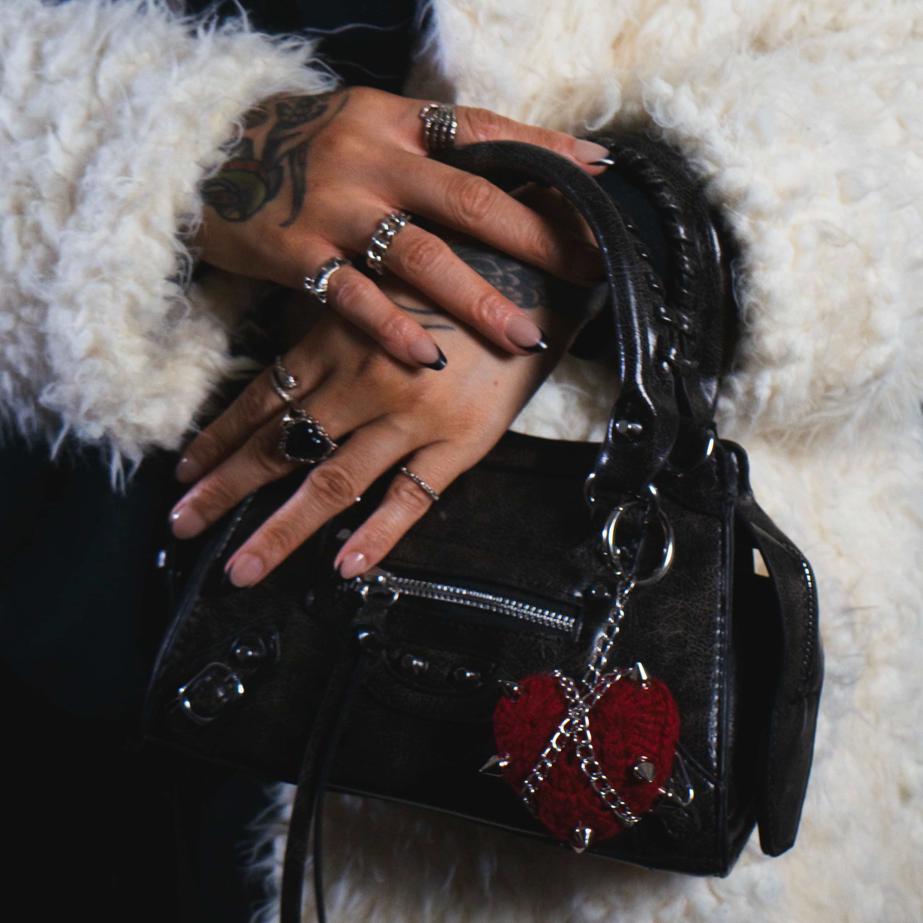 Black handbag with red heart charm held by a person wearing multiple rings on a white fur background