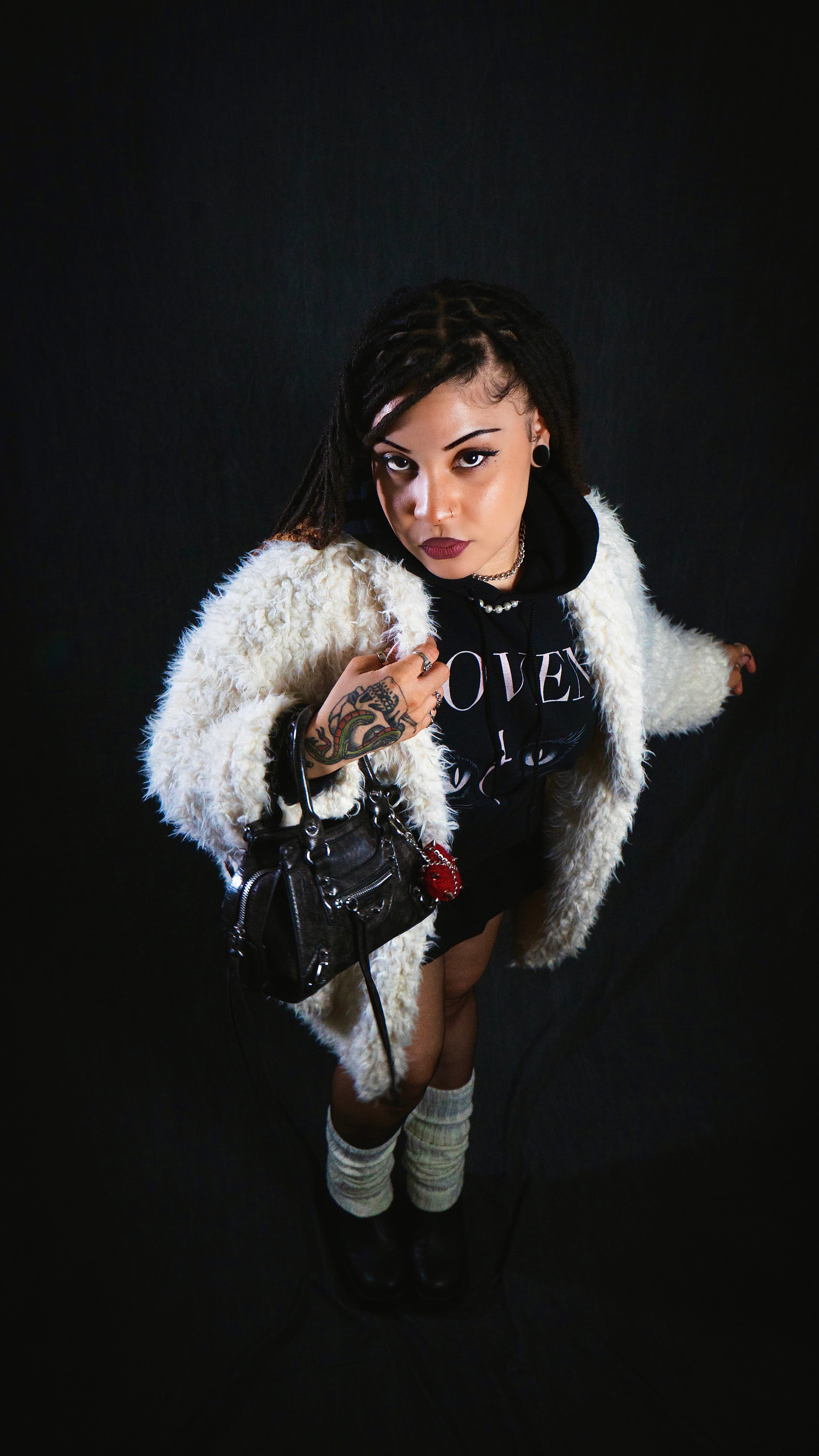 Person wearing a white fur coat and black outfit with 'COVEN' text, holding a black handbag against a dark background.