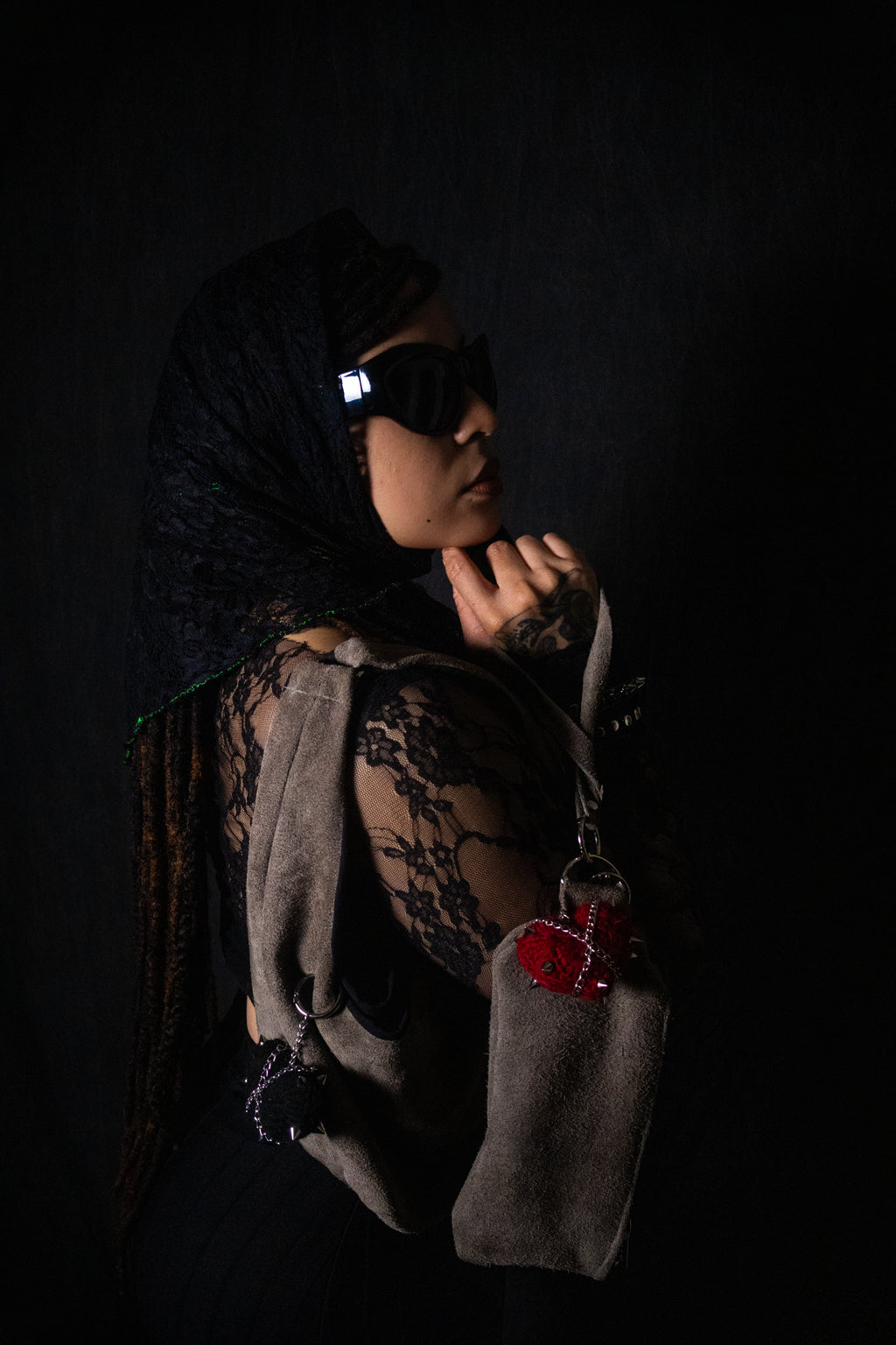 Person wearing a black head scarf and sunglasses, holding a gray bag with a red heart on a dark background