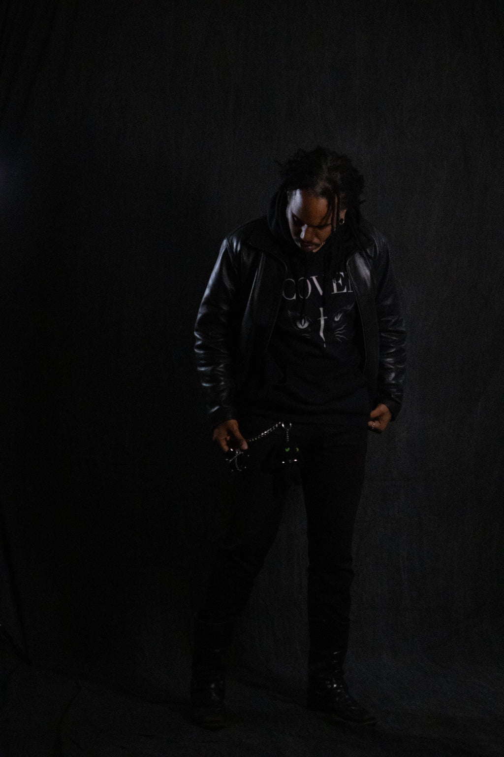 Person wearing a black leather jacket and hoodie with Coven text on it against a dark background