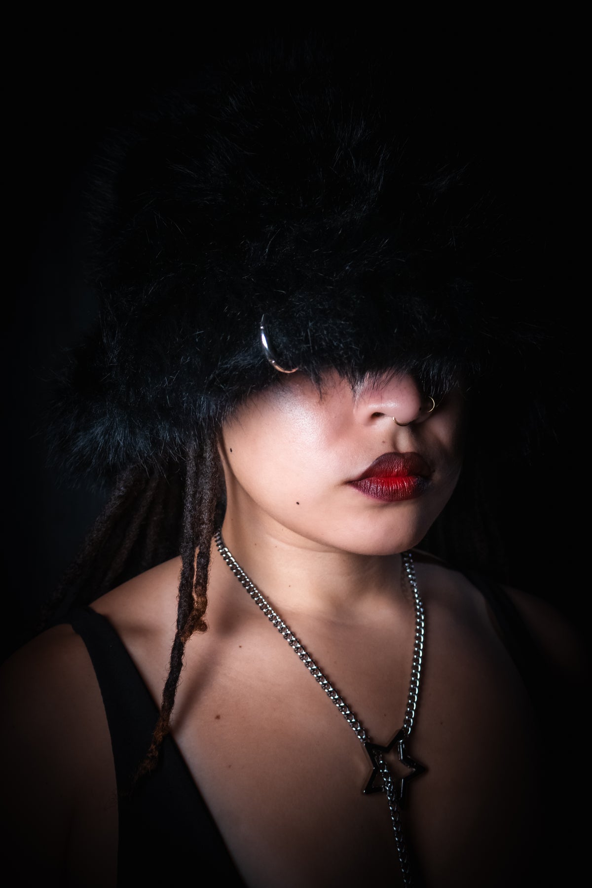 A close-up portrait of a person with dreadlocks wearing a black furry hat that obscures their eyes, a silver chain necklace with a star pendant, and red lipstick, set against a dark black background.