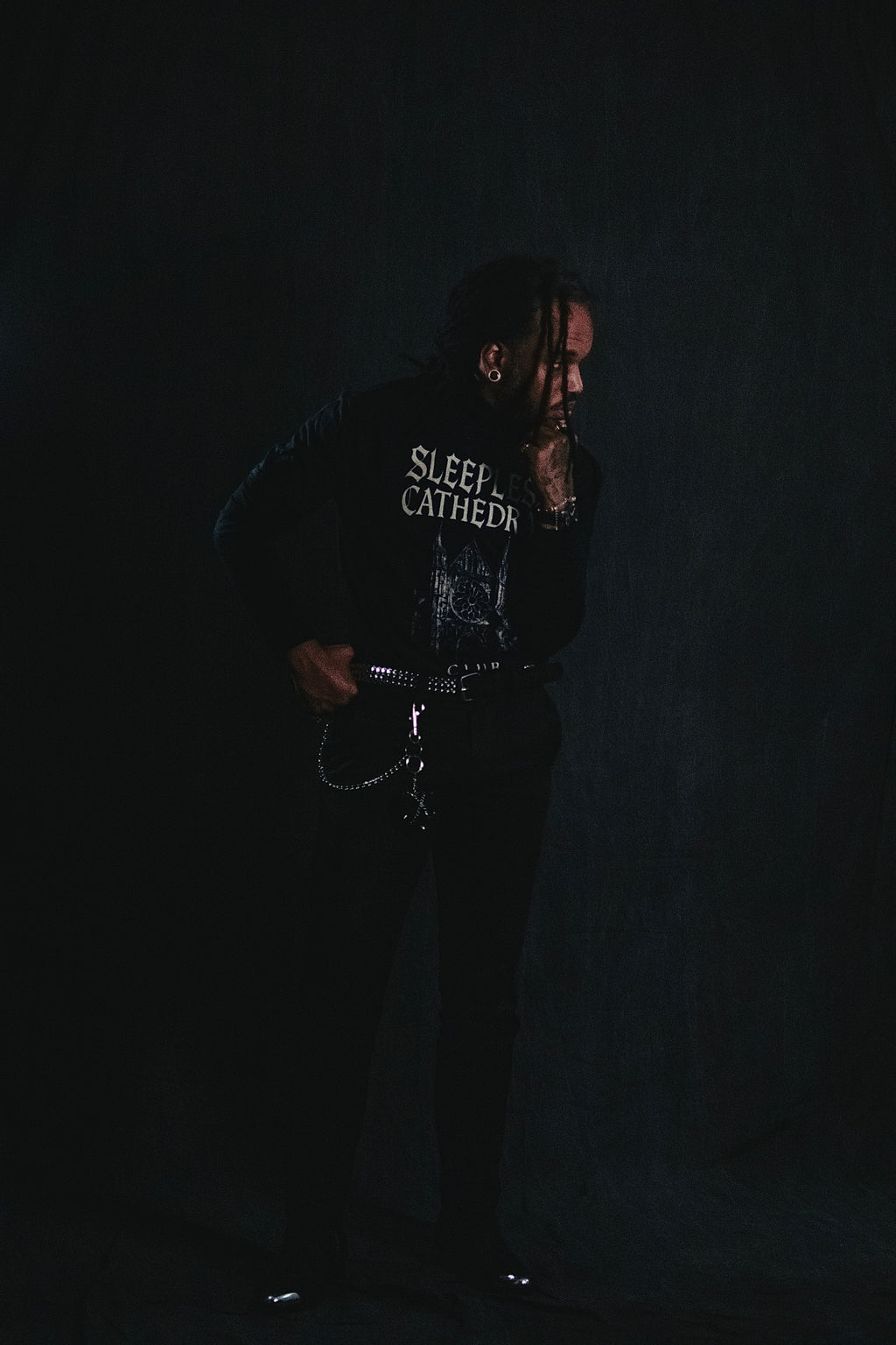Male model wearing the Coven x Coven ‘Sleepless Cathedral’ long sleeve shirt styled with black pants, studded belt, and chain accessories, standing against a dark gothic backdrop