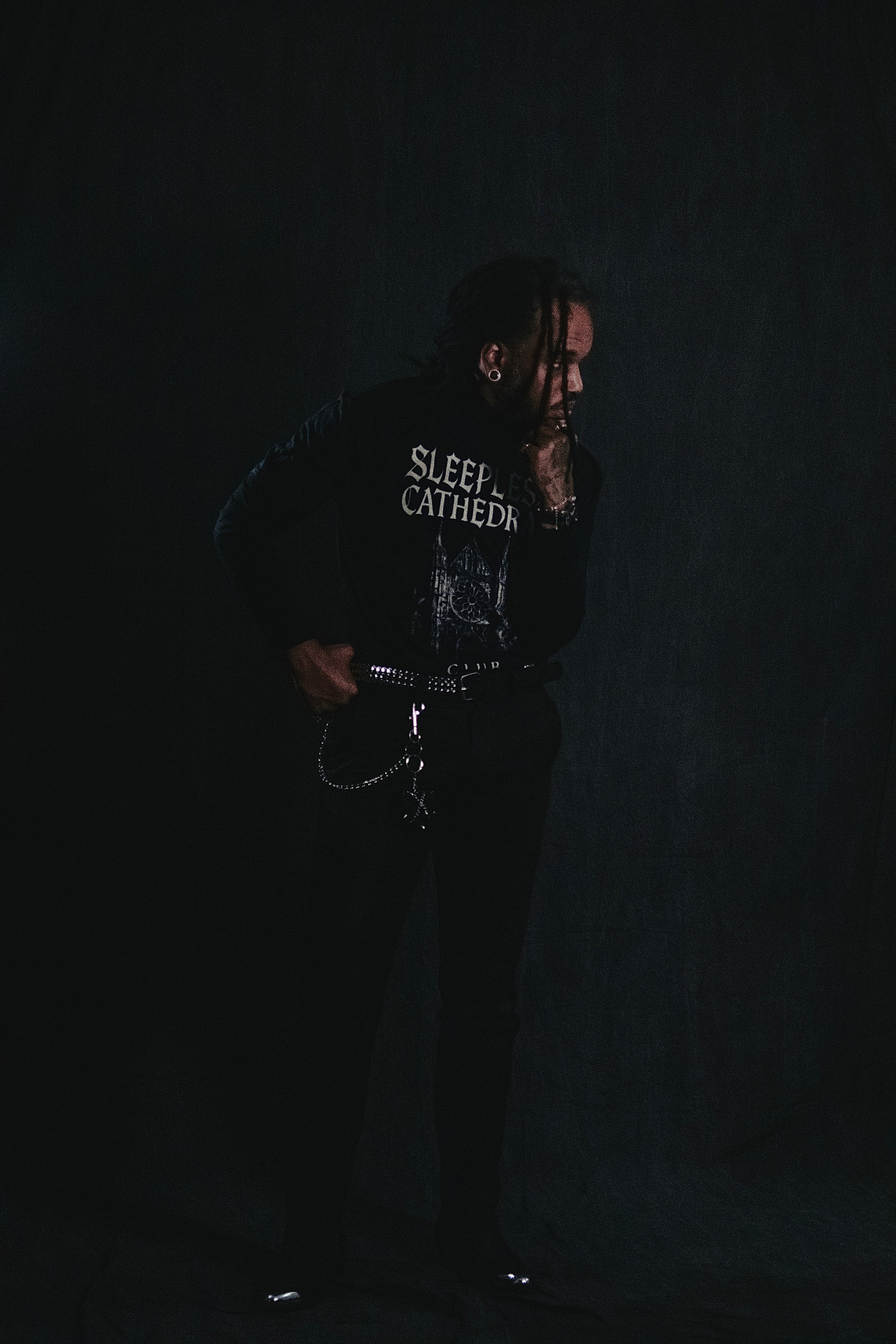 Male model wearing the Coven x Coven ‘Sleepless Cathedral’ long sleeve shirt styled with black pants, studded belt, and chain accessories, standing against a dark gothic backdrop